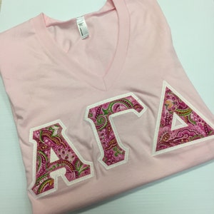 May include: A light pink V-neck t-shirt with the Greek letters ΑΓΔ in a paisley pattern. The letters are outlined in white. The shirt is folded neatly.