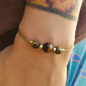May include: A beaded bracelet featuring three tiger's eye stones and gold-colored beads strung on a thin, tan cord. The bracelet is worn on a wrist with a colorful tattoo.