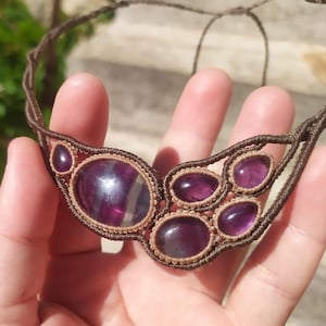 May include: A handmade macrame necklace featuring a series of oval and round purple gemstones. The gemstones are set in a tan macrame design with a brown cord. The necklace is held in a hand against a blurred green background.