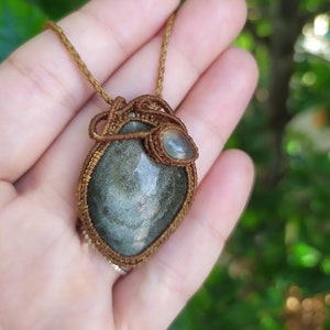 May include: A brown cord necklace with a green and grey stone pendant wrapped in brown cord. The stone is surrounded by a smaller, clear stone.