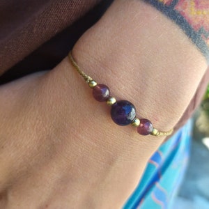 May include: A simple bracelet with a brown cord and purple beads. The bracelet has a larger purple bead in the centre.