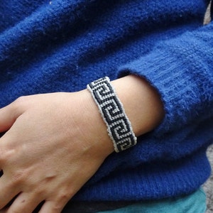 Può includere: Un braccialetto intrecciato bianco e nero con un motivo a chiave greca è indossato su un polso. Il braccialetto è fatto di piccoli fili fittamente intrecciati. La persona indossa un maglione blu e una maglietta turchese.
