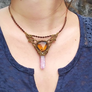 Op de afbeelding: Een bruine macramé ketting met een grote, heldere, oranje en bruine edelsteen hanger. De hanger is bevestigd aan een roze kristallen punt.