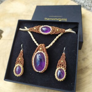 May include: A set of amethyst jewellery in a black box. The set includes a necklace, bracelet, and earrings. The jewellery is made with brown and gold cord and features amethyst stones.