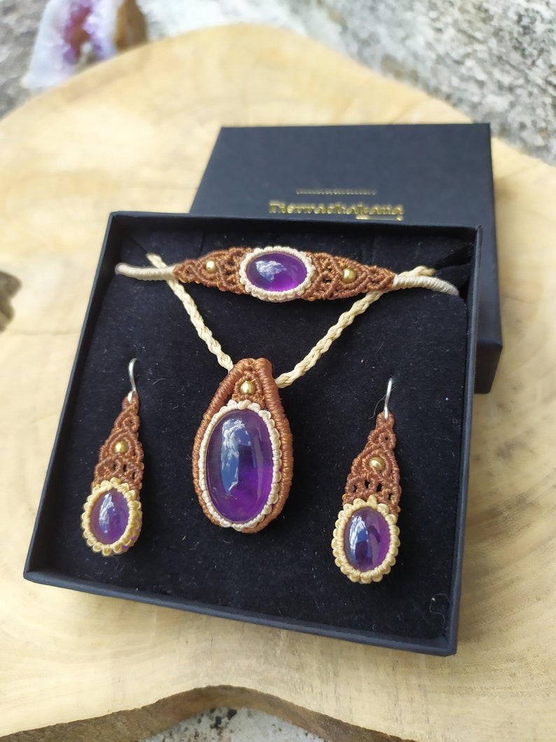 May include: A set of amethyst jewellery in a black box. The set includes a necklace, bracelet, and earrings. The jewellery is made with brown and gold cord and features amethyst stones.