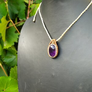 May include: A purple amethyst gemstone pendant necklace with a brown macrame cord and a white cord. The pendant is on a black mannequin.