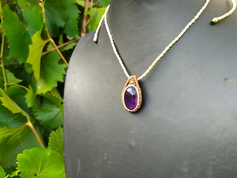 May include: A purple amethyst gemstone pendant necklace with a brown macrame cord and a white cord. The pendant is on a black mannequin.