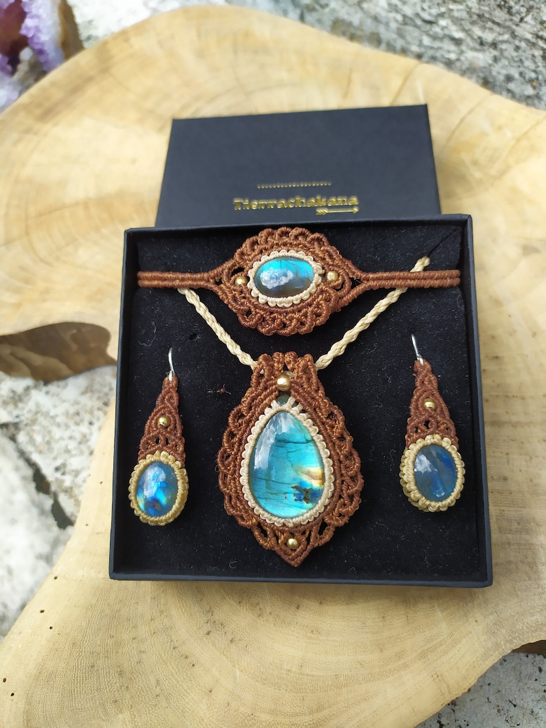May include: A brown macrame jewelry set with a blue labradorite gemstone pendant, earrings, and bracelet. The set is displayed in a black box with the text "Tierrachakana" on the lid.