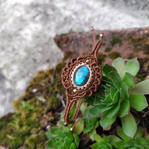 May include: A brown macrame bracelet with a blue labradorite gemstone in the center. The bracelet is tied with a knot and has gold beads on the ends.