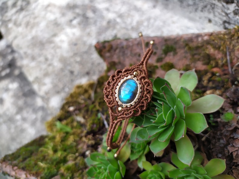 May include: A brown macrame bracelet with a blue labradorite gemstone in the center. The bracelet is tied with a knot and has gold beads on the ends.