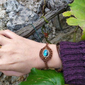 May include: A brown macrame bracelet with a blue labradorite gemstone. The bracelet is tied around a person's wrist.