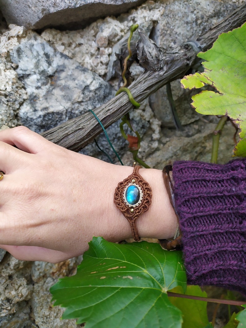 May include: A brown macrame bracelet with a blue labradorite gemstone. The bracelet is tied around a person's wrist.