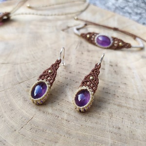 May include: A set of amethyst jewellery featuring a necklace, bracelet, and earrings. The jewellery is made with brown and cream macrame cord and features small gold beads. The amethyst stones are a deep purple colour.