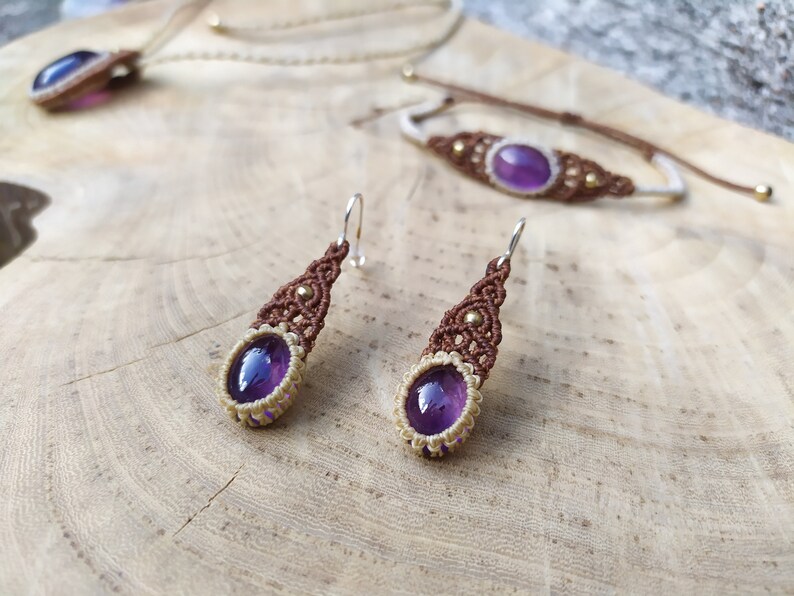 May include: A set of amethyst jewellery featuring a necklace, bracelet, and earrings. The jewellery is made with brown and cream macrame cord and features small gold beads. The amethyst stones are a deep purple colour.