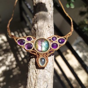 May include: A handmade macrame necklace featuring a large iridescent labradorite gemstone surrounded by smaller amethyst gemstones. The necklace is crafted with brown cord and has a delicate, intricate design.