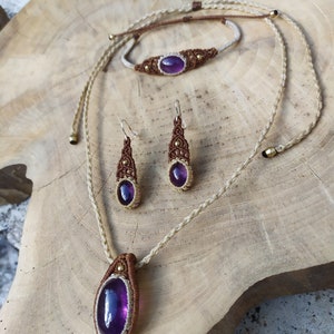 May include: A set of jewellery featuring a purple amethyst stone. The set includes a necklace, bracelet, and earrings. The jewellery is made with brown cord and gold accents.