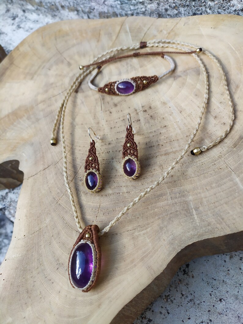 May include: A set of jewellery featuring a purple amethyst stone. The set includes a necklace, bracelet, and earrings. The jewellery is made with brown cord and gold accents.