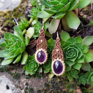 May include: A pair of brown macrame earrings with amethyst gemstones. The earrings are hanging from silver hooks and are accented with gold beads.