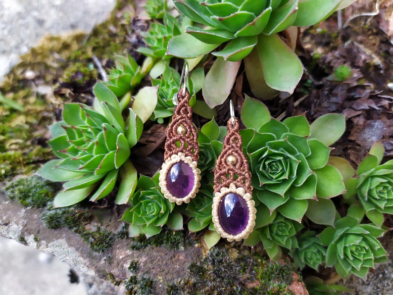 May include: A pair of brown macrame earrings with amethyst gemstones. The earrings are hanging from silver hooks and are accented with gold beads.