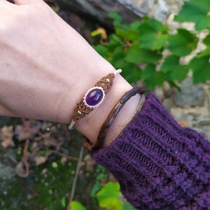 May include: A purple gemstone bracelet with brown and white macrame cord. The bracelet is tied with a knot and has a small gold bead.
