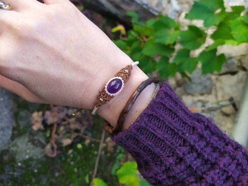 May include: A purple gemstone bracelet with brown and white macrame cord. The bracelet is tied with a knot and has a small gold bead.