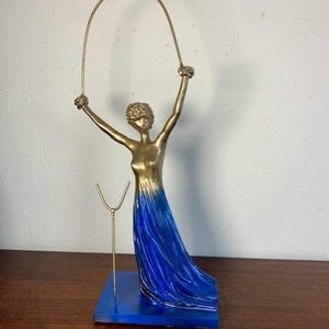 Può includere: Una scultura dorata di una donna che tiene una corda per saltare. L'abito della donna è un gradiente di blu e la scultura è montata su una base blu. Un piccolo supporto dorato è accanto alla donna. La scultura è su una superficie di legno.