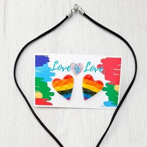 May include: A rainbow heart pendant necklace and matching earrings. The necklace is on a black cord and the pendant is a rainbow heart with a textured surface. The earrings are also rainbow hearts with a textured surface. The card says "Love is Love."