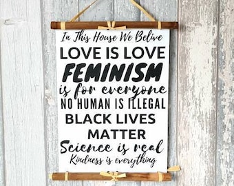 May include: A white rectangular wall hanging with the text "In This House We Believe, LOVE IS LOVE, FEMINISM is for everyone, NO HUMAN IS ILLEGAL, BLACK LIVES MATTER, Science is real, Kindness is everything." The sign is held by wooden dowels and a tan cord.