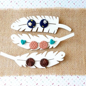 Puede incluir: Tres juegos de pendientes expuestos en soportes blancos con forma de pluma. El primer juego presenta pendientes florales azules y blancos, el segundo juego presenta pendientes verde turquesa y melocotón, y el tercer juego presenta pendientes de madera marrón.