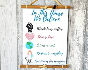 May include: A white scroll with the text "In This House We Believe" and five lines of text below it: "Black Lives Matter", "Love is Love", "Science is Real", "Kindness is Everything", and "Freedom is for Everyone". Each line of text is accompanied by a small graphic symbol.