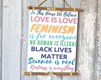 May include: A white sign with the text "In This House We Believe Love Is Love Feminism Is For Everyone No Human Is Illegal Black Lives Matter Science Is Real Kindness Is Everything" printed in rainbow colours.