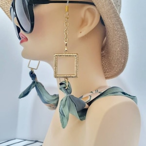 Puede incluir: Portagafas de cadena dorada con un colgante cuadrado. La cadena está unida a las gafas con lazos de silicona transparentes. El colgante es un cuadrado dorado con un borde texturizado. La cadena cuelga de la oreja de un maniquí que lleva un sombrero de paja y una bufanda verde.