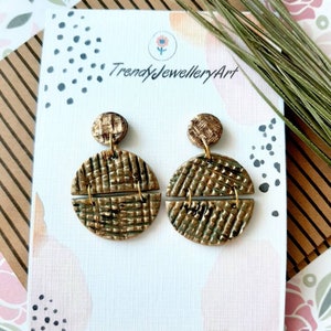 Op de afbeelding: Gouden en bruine geometrische oorringen met een geweven textuur. De oorringen worden getoond op een witte kaart met zwarte stippen en de tekst "Trendy Jewellery Art".