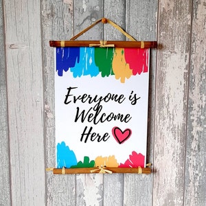 Könnte beinhalten: Ein weißes Schild mit dem Text "Everyone is Welcome Here" und einem roten Herzen. Das Schild hängt an einer Bambusrolle mit einem regenbogenfarbenen Hintergrund.