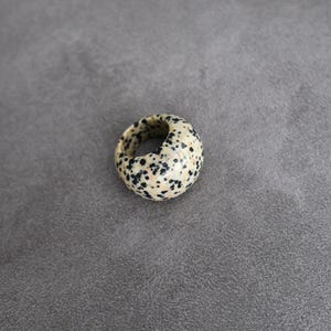 May include: A thick, rounded ring featuring a cream-coloured base with a dense pattern of small, irregular black spots throughout the surface. The band has a smooth, polished finish and a wide, chunky profile that creates a continuous circular shape.
