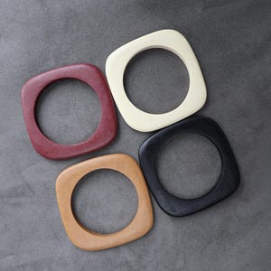 May include: Four square-shaped bangles arranged in a cluster. The set includes one deep red, one cream, one light tan, and one black bangle. Each piece features a smooth, rounded outer edge and a circular inner opening, crafted from a solid material.