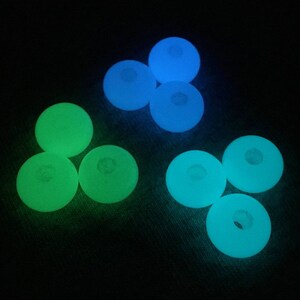 May include: Nine glow-in-the-dark round beads, some green and some blue, arranged in a triangular pattern on a dark surface.
