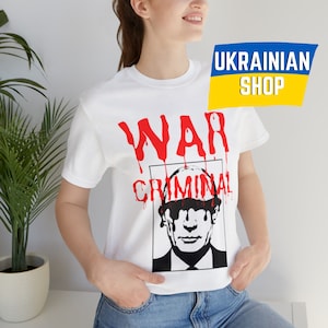 May include: White t-shirt with a black and white image of a man's face with the word "WAR" in red dripping paint above the face and the word "CRIMINAL" in red dripping paint below the face. The text "UKRAINIAN SHOP" is in the upper right corner of the image.