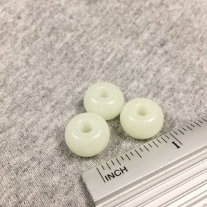 May include: Three white glow-in-the-dark round beads with a hole in the center. The beads are about 1 inch in diameter and are sitting on a gray fabric surface next to a ruler.