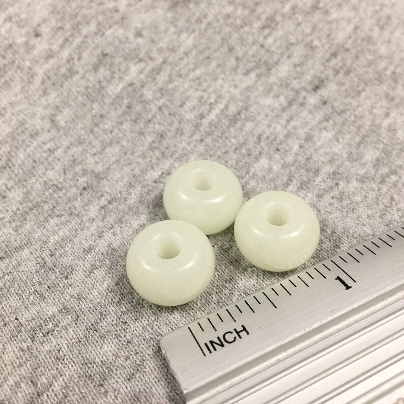 May include: Three white glow-in-the-dark round beads with a hole in the center. The beads are about 1 inch in diameter and are sitting on a gray fabric surface next to a ruler.