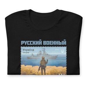 May include: Black t-shirt featuring a postage stamp design with the text "Русский военный" and "Україна". The stamp depicts a warship and a soldier. The year "2022" is also visible. The shirt is folded.