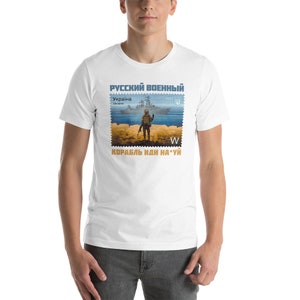 May include: White t-shirt featuring a stamp design with a soldier, a warship, and text in Cyrillic script. The stamp design includes the text "RUSSIAN MILITARY" and "UKRAINE". The shirt also has the text "SHIP GO F*** YOU".