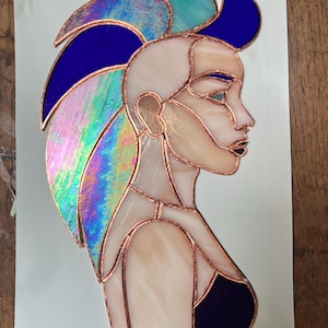 May include: A stained glass panel depicting a profile of a person with a mohawk hairstyle. The piece features iridescent blue, teal, and clear glass segments joined by copper foil lines, showcasing a side view of a face and upper torso.