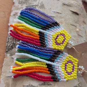 May include: A pair of beaded earrings with a rainbow fringe and a yellow and purple symbol. The earrings are hanging from a birch bark background.