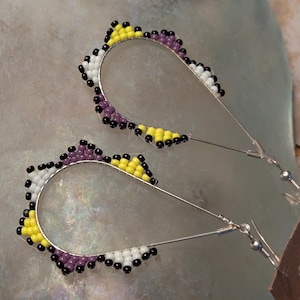 May include: A pair of handcrafted earrings with a teardrop shape. The earrings feature a silver wire frame adorned with a beaded design in white, yellow, purple, and black. The earrings are displayed on a light gray surface.
