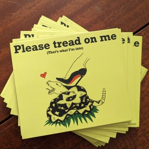 Puede incluir: Una pila de tarjetas amarillas con el texto "Please tread on me (That's what I'm into)" y una serpiente en blanco y negro con un corazón rojo y un zapato de tacón negro pisándola.