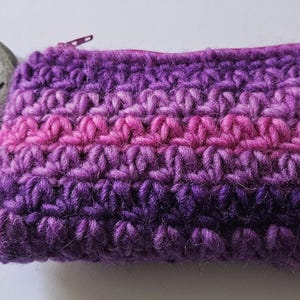 May include: A rectangular crochet pouch featuring a gradient of purple and pink yarn with a visible zipper closure. Beside the pouch sits a smooth, round, light gray stone with the word love written in black cursive script on its surface.