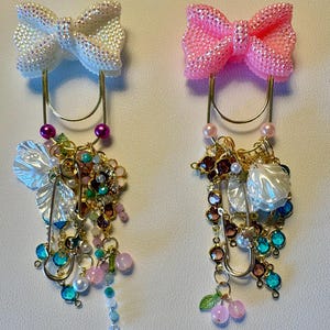 May include: Two decorative bow-shaped paper clips. One is white with iridescent glitter, the other is pink. Both have gold-colored metal clips and are adorned with dangling charms, beads, and faux pearls in various colors.