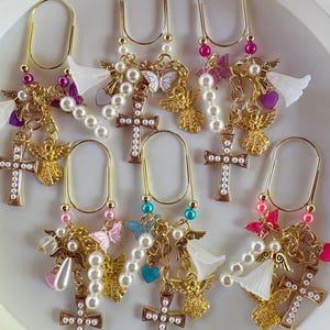 May include: A collection of six gold-toned metal paper clips, each adorned with dangling charms including white faux pearls, gold-toned angel figures, small colorful butterflies, heart-shaped beads, and crosses inlaid with small white faux pearls.