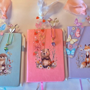 May include: Five colorful journals with animal illustrations and decorative bookmarks. Each journal features a different animal: a squirrel, otter, bear, fox, and bird. The journals have matching pens and decorative ribbons.